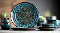 Handcrafted ceramic dinnerware set in turquoise and black with spiral pattern design, featuring plates, bowls and cups against dark background with succulent decor.