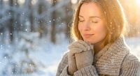Young Caucasian woman in cozy knit sweater and mittens enjoying peaceful winter moment with closed eyes as snowflakes fall in bright sunlight outdoors.