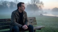 Thoughtful middle-aged Caucasian man in olive jacket sitting on wooden bench during misty sunrise, contemplating by lake in peaceful park setting.