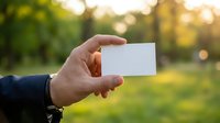 Male hand holding blank white business card outdoors in green park with bokeh background, mockup for branding and design.