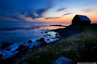 Silhouette of coastal barn at dusk with dramatic sky, rocky shore, and wildflowers in foreground. Long exposure creates misty effect on ocean waves against blue twilight.