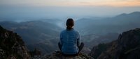 Young adult female with dark hair, wearing blue jacket, sitting on mountain cliff edge overlooking misty valley landscape at sunrise or sunset. Solitude and contemplation concept for wellness and travel.