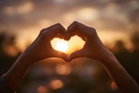 Hands forming heart shape silhouette against warm golden sunset sky with bokeh lights creating romantic atmosphere for love and connection concepts.
