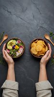 Female hands holding wooden bowls with fresh salad and crispy potato chips on dark slate background, styled for healthy eating and snack food choices.