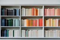 Books arranged by color on white wooden shelves creating rainbow gradient effect from dark blue through orange to pink, modern minimalist library interior.