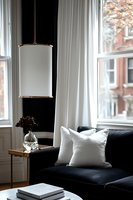 Modern living room corner with black velvet sofa, white throw pillows, and cylindrical pendant lamp. Minimalist interior design features marble coffee table and dried flowers in vase.