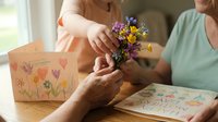 Elderly woman receiving small wildflower bouquet from caregiver with handmade greeting cards on table, showing intergenerational care and appreciation.
