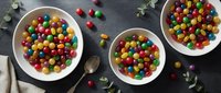 Colorful round candies in white bowls on dark surface with eucalyptus leaves and scattered sweets for festive celebrations and party treats.