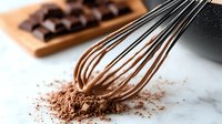 Chocolate whisk with cocoa powder on marble surface, dark chocolate pieces on wooden board in background, close up view for baking and dessert preparation.