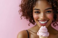 Young woman with curly brown hair and medium skin tone holding a pink frosted cupcake against pink background, smiling brightly for bakery, dessert, and food advertising campaigns.