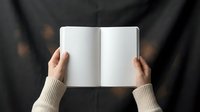 Hands holding open blank notebook with empty white pages on dark textured surface. Top view of person in beige sweater reading journal for writing notes.