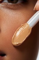 Makeup artist applying liquid foundation on African American woman's face with cosmetic spatula, showing texture of product and glowing skin. Professional beauty care.