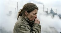 Middle-aged Caucasian woman coughing in polluted industrial area with factory smokestacks emitting toxic fumes in background for health awareness campaigns.