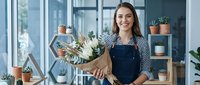 Young woman with long dark hair and light skin tone, wearing a dark apron, holding a bouquet of white and green flowers in a bright, modern flower shop with potted plants and shelving. Florist, small business owner, retail environment.