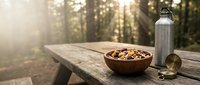 Wooden bowl filled with mixed nuts and dried fruits on rustic table outdoors with water bottle, healthy snacking concept for camping adventures.