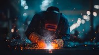 Welder in protective mask working with metal, creating bright orange sparks against dark blue industrial background with bokeh lights.
