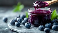 Fresh blueberry jam being spooned from glass jar with wooden spoon, surrounded by ripe berries and mint leaves on rustic stone surface for cooking.