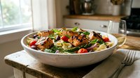 Fresh Mediterranean salad with grilled chicken, mixed greens, cherry tomatoes and feta cheese in white bowl on rustic wooden table in bright kitchen.