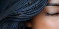 Close up of Asian woman with closed eyes and long dark blue hair flowing in wind, showing natural makeup and serene expression. Partial face portrait on dark background.