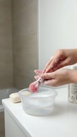 Woman hands washing pink makeup brush in glass bowl with soapy water on bathroom counter for beauty tool cleaning and hygiene maintenance routine.