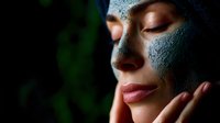 Young woman with clay face mask enjoying spa treatment against dark background with green plant elements. Wellness and skincare concept.