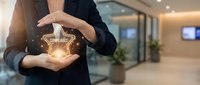 Professional businesswoman in dark suit holding glowing compliance shield symbol with protective hands in modern corporate office environment.