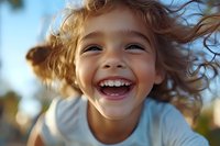 Joyful young girl with curly hair laughing outdoors in natural sunlight, showing genuine happiness with bright smile and windswept hair against blurred background.