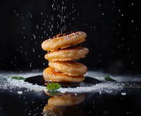 Stack of glazed donuts with falling sugar powder on black reflective surface, dramatic dark food photography with mint leaves and moody lighting.