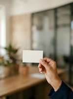 Hand holding blank white business card in modern office environment with blurred background, suitable for branding, corporate identity, and marketing mockup designs.