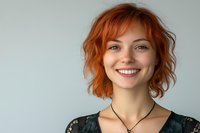 Young Caucasian woman with vibrant red wavy bob haircut and natural makeup smiling warmly at camera against light background. Genuine happiness expression.