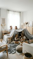 Happy family building blanket fort together in cozy living room with children playing underneath makeshift tent made from chairs and sheets.