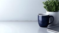 Dark blue speckled ceramic mug on white surface next to green plant in pot, minimalist kitchen decor setup for morning coffee or tea routine.