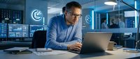 Focused businessman working late on laptop in modern office with data screens and blue lighting for corporate technology projects.