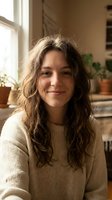 Young woman with curly brown hair smiling warmly in cozy home interior with plants and natural light streaming through window wearing beige sweater.