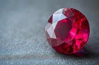 Brilliant cut round ruby gemstone with deep red color and faceted surface on gray textured background, showcasing luxury jewelry craftsmanship and precious stone detail.