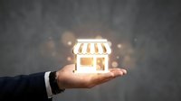 Businessman hand holding glowing storefront icon with striped awning against textured gray background, representing retail business startup and e-commerce opportunity.