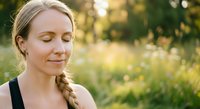 Caucasian woman with blonde braided hair wearing black tank top, eyes closed with serene expression in sunlit meadow with green foliage background for wellness and lifestyle content.