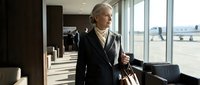 Senior Caucasian businesswoman in elegant suit walking through modern airport terminal with luggage, professional travel concept for corporate mobility.
