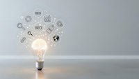 Glowing light bulb with SEO marketing icons floating above on gray background representing digital strategy and search engine optimization concepts.