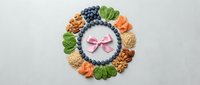Healthy snack platter with nuts, berries, carrots and greens arranged in circle around yogurt bowl with pink bow for nutritious eating and wellness lifestyle.