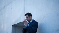 Young Caucasian businessman in suit praying with hands together, eyes closed, standing against concrete wall outdoors.