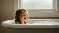 Young girl with curly hair enjoying bubble bath in white bathtub by window, childhood hygiene and self-care routine in bright bathroom interior.