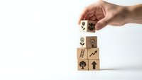 Hand placing wooden cube with lightbulb icon on stack showing business growth symbols including ladder, arrow, tree on white background.