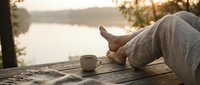 Relaxed person enjoying peaceful morning coffee by lake during golden hour sunrise. Cozy lifestyle moment with warm blanket and serene waterfront view.