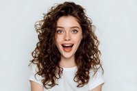 Young woman with curly brown hair and bright eyes expressing excitement and joy against white background, wearing casual white t-shirt.