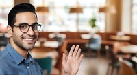 Young South Asian male professional wearing glasses and blue shirt waving hello in modern office environment, suitable for business, corporate communications, and workplace diversity concepts.