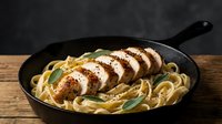 Sliced grilled chicken breast with creamy pasta in cast iron skillet on wooden table. Restaurant quality meal with herbs and sauce for food photography.