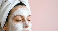 Young woman with white towel on head applying facial mask for skincare routine and spa treatment at home on pink background.