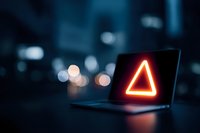 Laptop with glowing red triangle warning sign against dark blurred city background, concept of cybersecurity alert or system error.