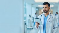 Young Hispanic male doctor with stethoscope holding smartphone in modern hospital corridor, smiling confidently while wearing white medical coat.
